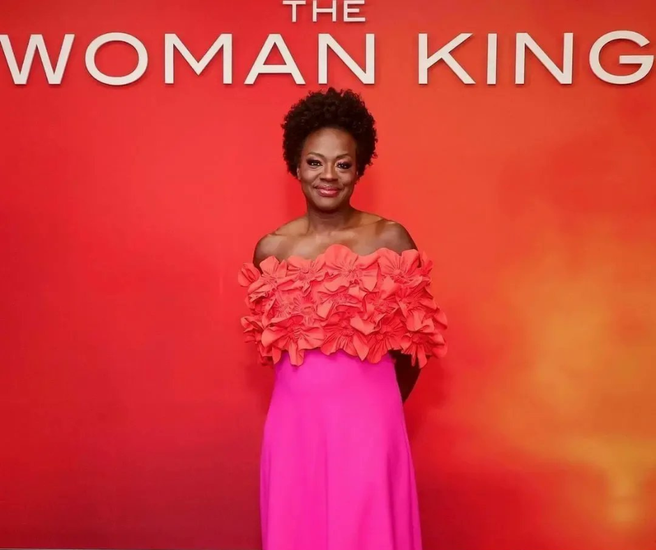 Actress Viola Davis in red carpet attire in front of The Woman King background